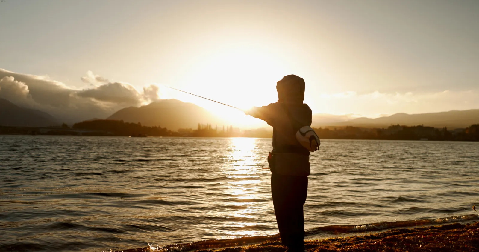 Angler casting from a lakeshore below mountains at sunset